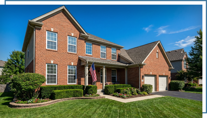 Traditional brick house with picture windows, manicured lawn, and American flag, showcasing natural light and outdoor views, ideal for Kansas City homeowners seeking window replacement options.